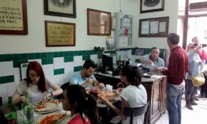 Wnętrze najstarszej pizzerii w Neapolu. Ludzie jedzą pizzę, a za ladą obsługuje kelner.