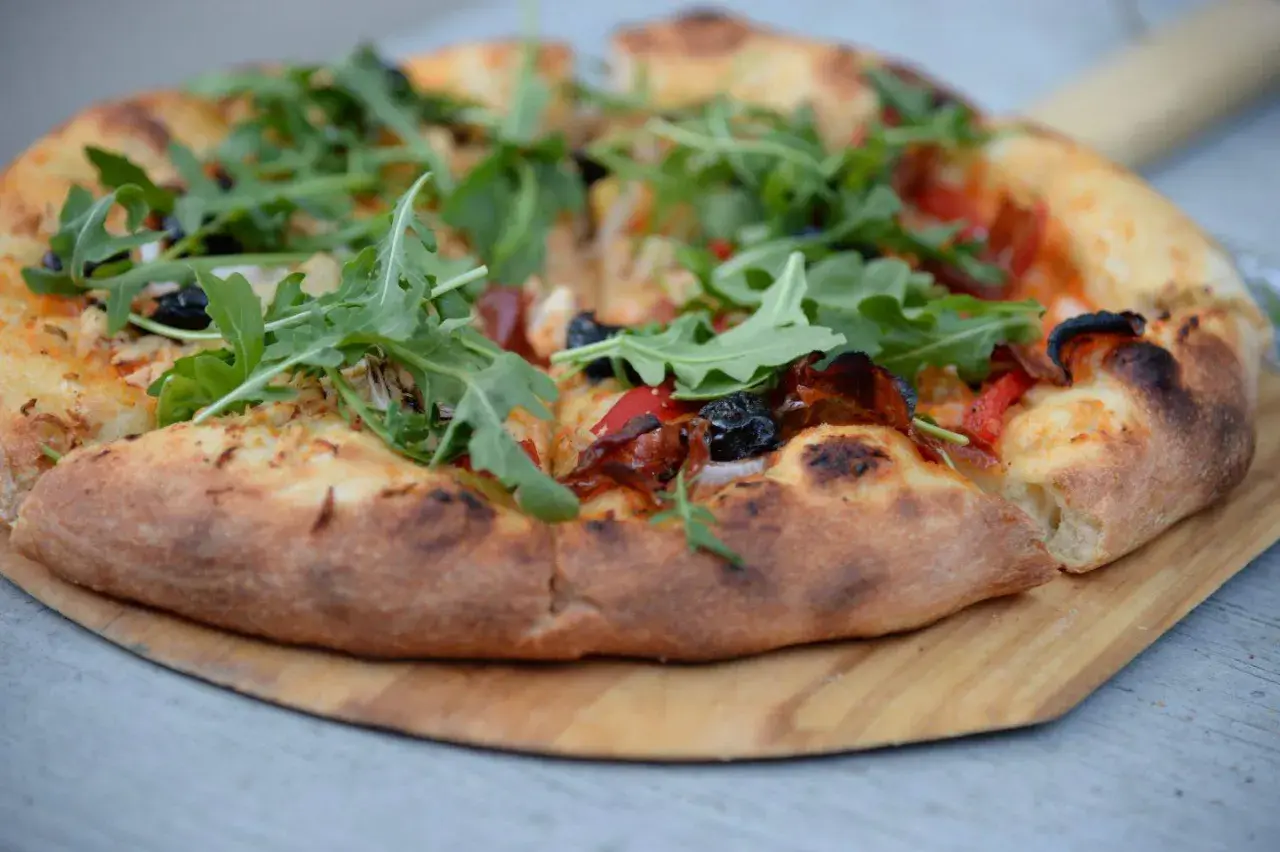 Puszyste ciasto pizzy z rukolą, oliwkami i papryką, podana na drewnianej desce.