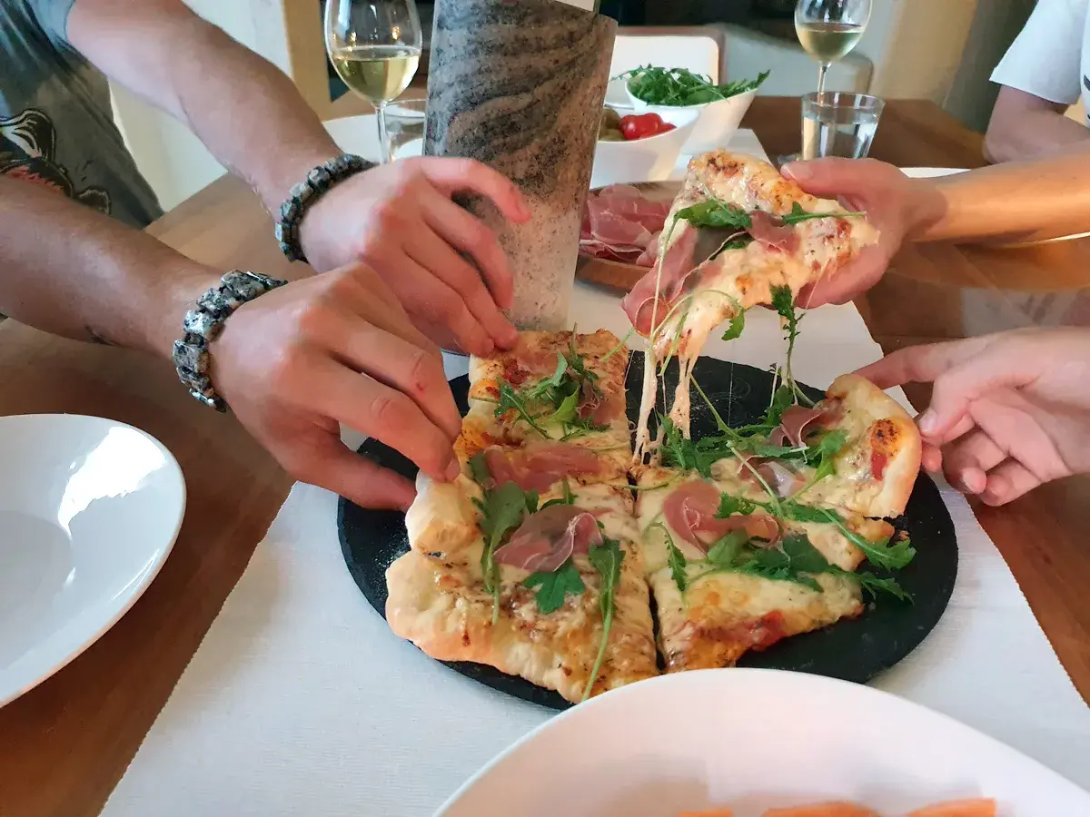 Ręce sięgają po kawałki pysznej pizzy na kamieniu w piekarniku, ciągnący się ser i rukola.