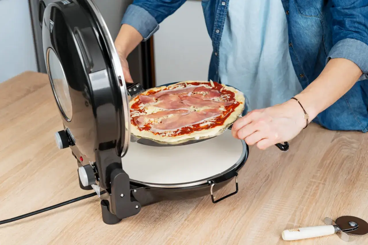 Osoba wkłada pizzę z szynką do pieca. Zastanawiasz się, ile powinna się piec pizza? Ten piec pomoże Ci to ustalić.