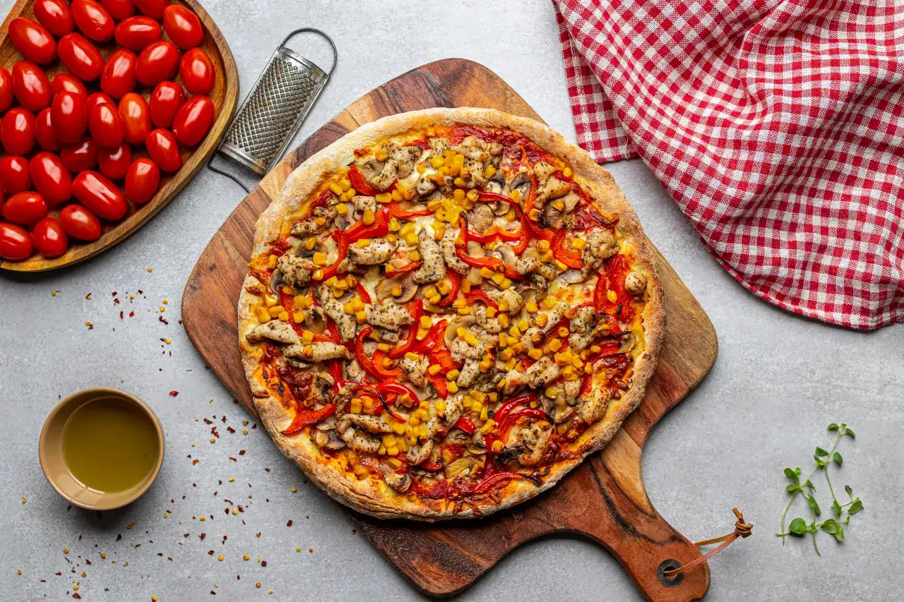 Pyszna pizza z kurczakiem, kukurydzą i papryką, gotowa do podania. Obok miseczka z pomidorkami i oliwą.