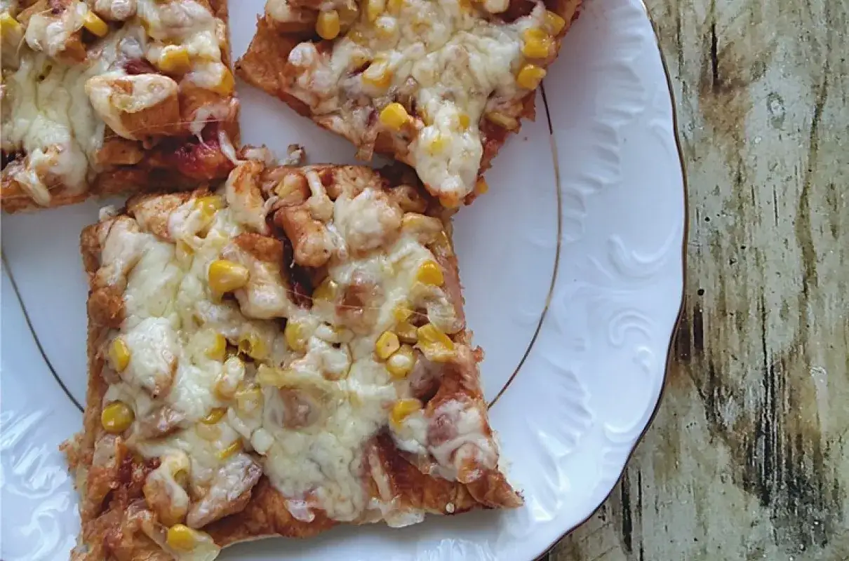 Kawałki pysznej pizzy z kurczakiem i kukurydzą, zapieczone pod ciągnącym się serem, podane na białym talerzu.