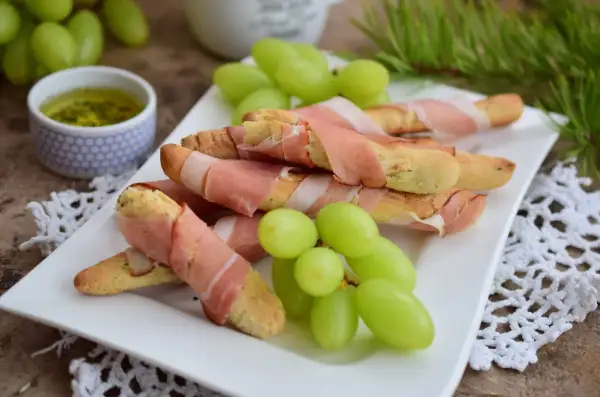 Grissini z szynką parmeńską, zielone winogrona i sos na talerzu. Idealna przekąska na przyjęcie.